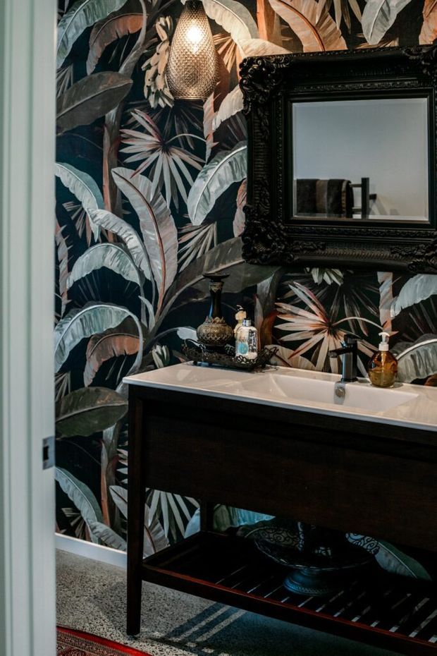 A richly styled bathroom vanity area featuring dramatic tropical botanical wallpaper, a dark timber console-style vanity with a white integrated basin, an ornate black-framed mirror, patterned accessories, and a woven pendant light. The palette is dark, warm, and highly decorative.