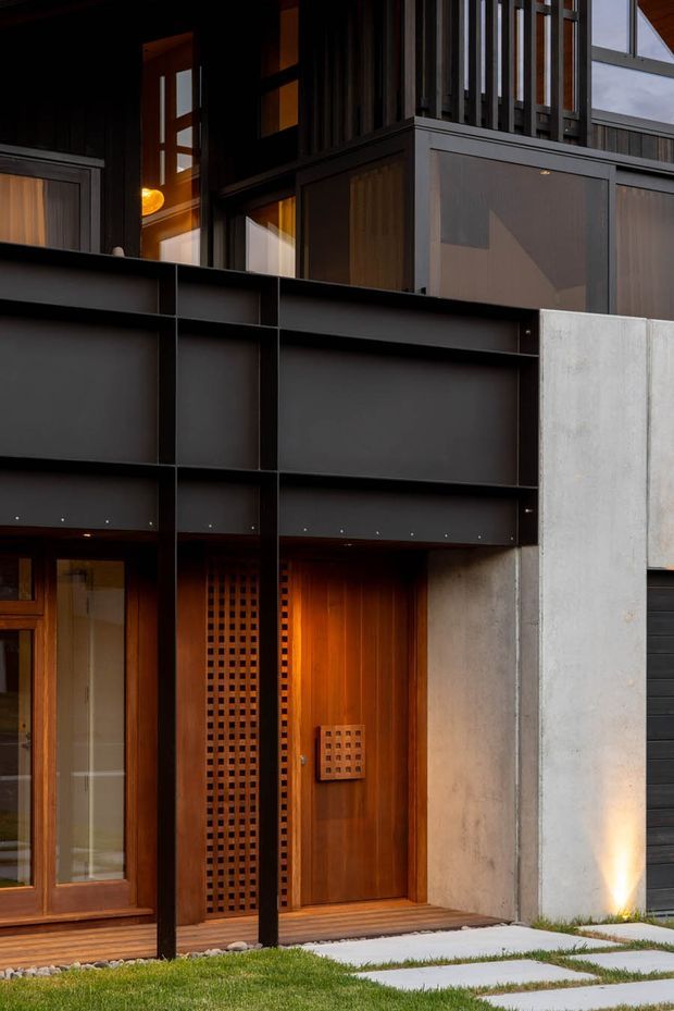 Close-up exterior view of a contemporary residential entrance. The facade combines dark metal framing and panels, warm timber glazing and a timber front door, vertical slatted screening, perforated timber detailing, and a light raw concrete wall. Low horizontal canopy elements and integrated warm lighting create a dramatic, refined entry sequence, with stepping-stone paving and lawn in the foreground.