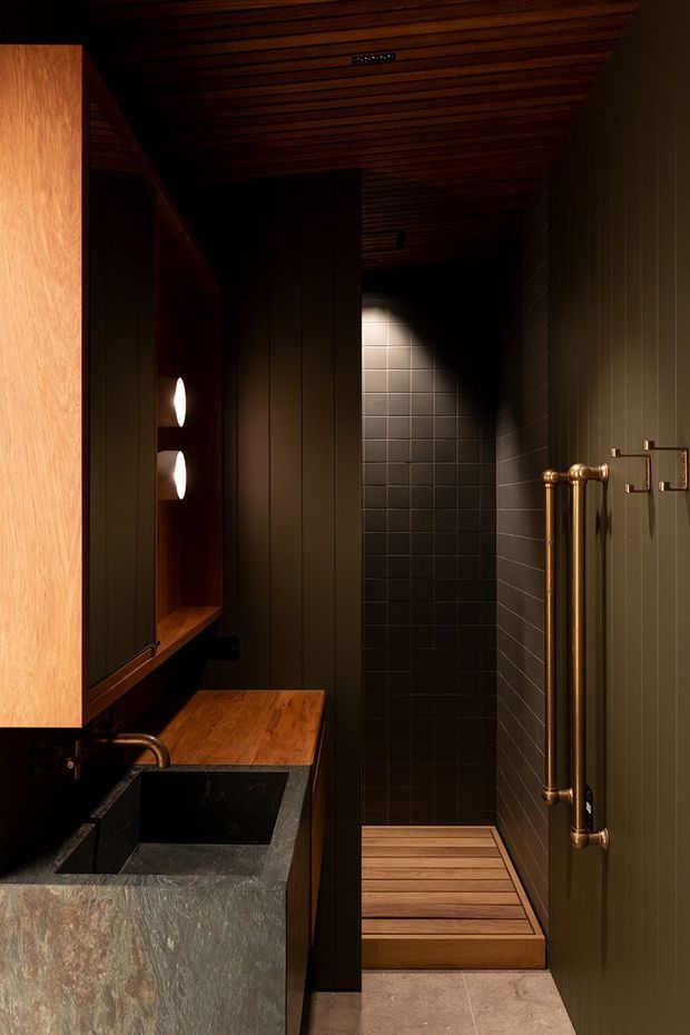 Moody, narrow contemporary bathroom with a dark green vertically panelled wall, dark tiled shower area, timber slatted ceiling, timber vanity surface, and a substantial grey stone basin. A tall brass heated towel rail and brass hardware add warmth. The shower floor includes a raised timber slat platform, and concealed lighting creates a dramatic atmosphere.
