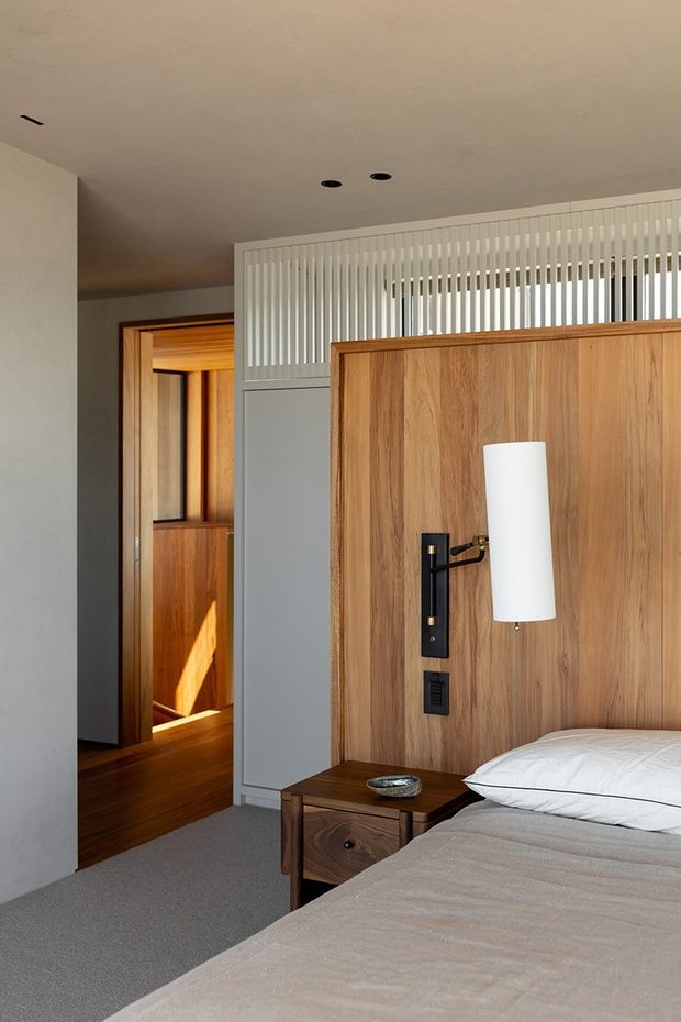 A warm, contemporary bedroom interior with a partially visible bed in the foreground, a timber feature headboard and integrated bedside table, and a slim wall-mounted reading light. A timber-lined doorway leads to an adjacent warm wood-lined space, while a white vertical-slat screen provides visual separation near the ceiling. Neutral carpet, plaster walls, recessed ceiling lights, and natural timber finishes create a refined, softly lit atmosphere.