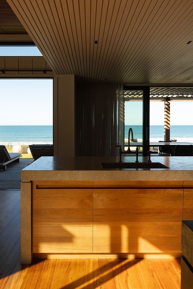 A warm, coastal kitchen interior centered on a substantial timber island with a dark stone benchtop, integrated sink and sculptural faucet. The space opens directly toward a bright beach and ocean through extensive floor-to-ceiling glazing. Timber slatted ceiling, dark framed glazing, timber flooring, and an outdoor dining terrace create a strong indoor-outdoor connection.
