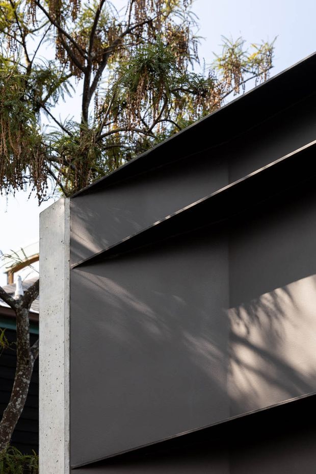 Close-up exterior architectural detail showing a dark charcoal, sharply angled folded roof or wall plane meeting a raw concrete vertical edge. The minimalist geometric facade is set against leafy vegetation, with strong sunlight and tree shadows across the dark surface.