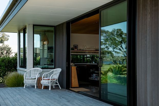 Contemporary residential exterior showing a covered deck or terrace with grey timber-look flooring, dark vertical cladding, black-framed sliding glass doors, and a partially visible interior bar or living area. White outdoor lounge chairs sit beside a glazed corner, with landscaped greenery reflected in the glazing.