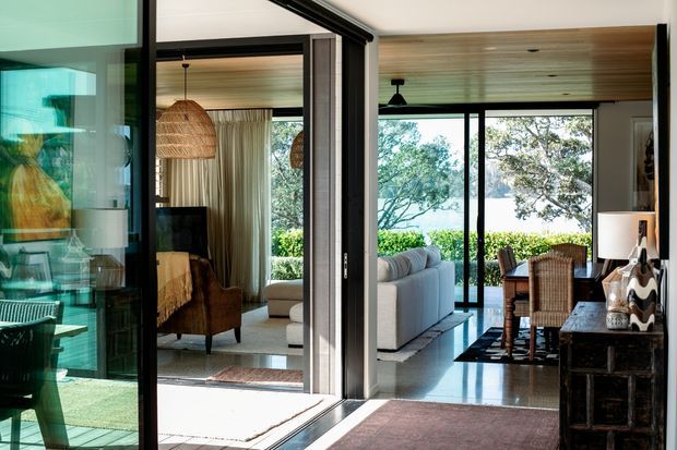 Interior view through black-framed glazed partitions and sliding doors, showing an open-plan living and dining area with extensive views to lush landscaping and water beyond. The space features a timber-lined ceiling, polished light flooring, neutral upholstered seating, woven pendant lighting, a wicker dining set, and dark built-in joinery. Strong indoor-outdoor connection and abundant natural light define the scene.