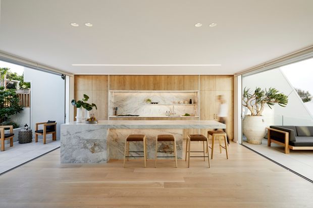Bright open-plan kitchen and entertaining space with a large marble-clad island, timber cabinetry and integrated shelving, pale timber flooring, and full-height corner glazing opening to outdoor areas. The room includes bar seating and a small lounge area with indoor plants and restrained contemporary furnishings.