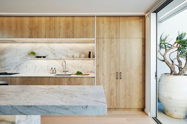 A refined contemporary kitchen with full-height light timber cabinetry, open shelving, and a dramatic veined stone splashback and island benchtop. A stainless-steel sink and tap sit beneath the shelving, while large sliding glass doors open to a balcony or terrace with a potted plant. The palette is warm and natural with clean-lined built-in joinery.