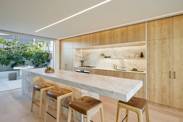 A bright, spacious contemporary kitchen featuring a large waterfall-edge stone island with four leather-upholstered stools, full-height light timber cabinetry, and a marble-look stone splashback with open display shelving. Integrated appliances and minimal linear lighting create a clean, refined interior, while glazing and indoor greenery bring in natural light.
