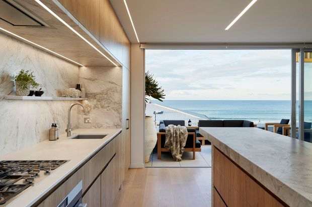 A refined open-plan kitchen overlooking the ocean, with a large stone island and waterfall benchtop, integrated timber cabinetry, marble-look wall surfaces, gas cooktop, and a dining/living area opening onto a seaside terrace. Linear recessed lighting and warm timber finishes create a clean contemporary coastal interior.