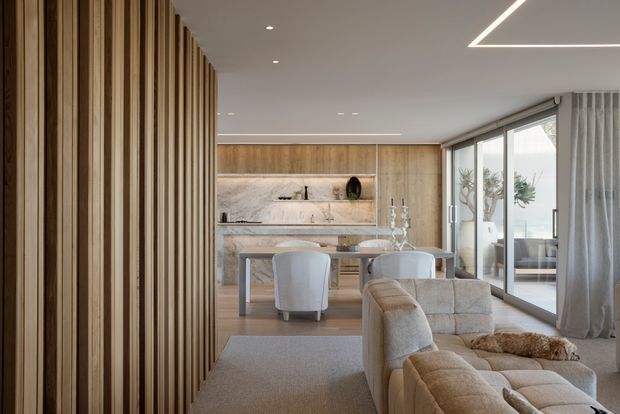 A refined open-plan living and dining area with a warm timber feature screen at left, a light wood and marble kitchen or bar wall at the rear, a round dining table, upholstered dining chairs, and a large neutral modular sofa. Full-height sliding glass doors open to an outdoor area, while linear recessed ceiling lighting and soft curtains create a calm, contemporary interior.