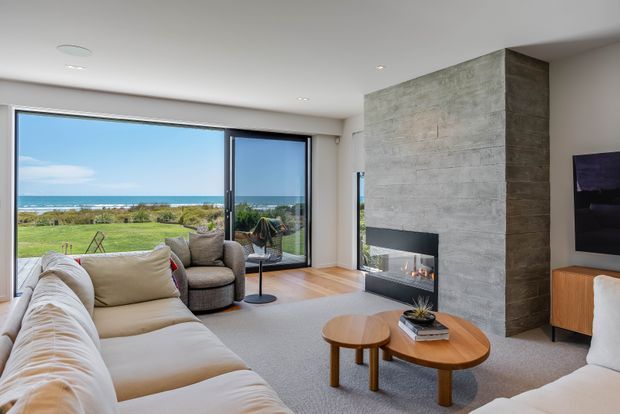 Bright contemporary living room with a large cream sectional sofa, rounded timber coffee tables, a grey lounge chair, and a wall-mounted television. A substantial textured grey concrete or stone fireplace feature wall anchors the room, with a linear fireplace inset. Full-height glazing and a sliding glass door open to a lawn and ocean view, creating a light coastal outlook.