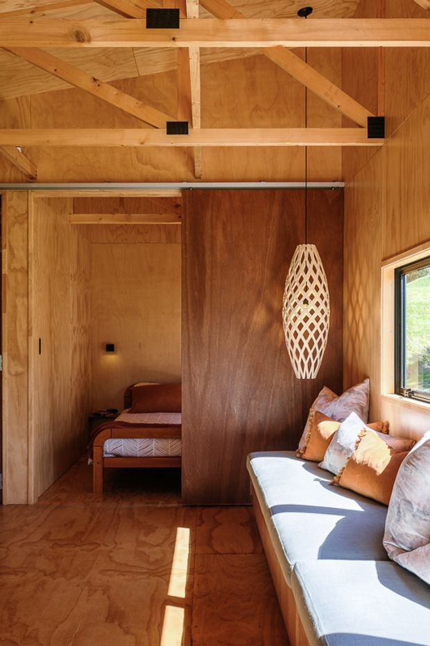 A compact, warm timber-lined interior with exposed plywood walls, ceiling, flooring, and structural rafters. The main space contains a built-in window seat or sofa with cushions, a woven pendant light, and a partially enclosed sleeping nook with a bed visible through an opening. Natural light enters through the side window, creating a rustic cabin-like atmosphere.
