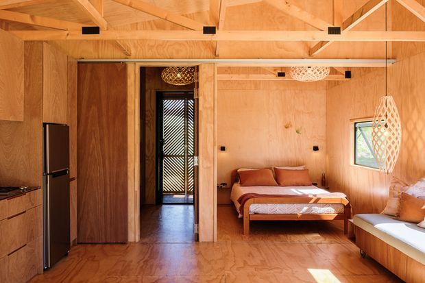 Warm, compact open-plan timber-lined interior combining a kitchenette and sleeping area. The room features exposed plywood wall and ceiling panels, timber floor and ceiling beams, built-in wood cabinetry, a tall dark refrigerator, a double bed, and a built-in window seat. A doorway leads to a narrow hall or exterior transition, while patterned pendant lights and a small window add detail and natural light.