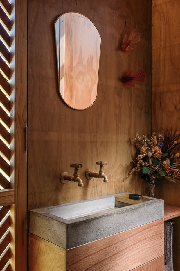 A warm, richly detailed bathroom vanity featuring a rectangular concrete trough basin on timber cabinetry, exposed vintage-style wall-mounted taps, and a rounded rectangular mirror set into dark timber wall panelling. Decorative dried flowers and a towel add styling, while a louvred window brings in filtered light.
