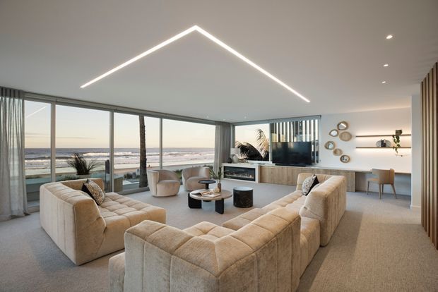 A spacious contemporary living room with a large cream modular sectional arranged around a low coffee table and a built-in fireplace/media unit. Full-height glazing overlooks a beach and ocean, while recessed spotlights, a geometric linear ceiling light, pale timber joinery, and neutral textured finishes create a refined coastal atmosphere.