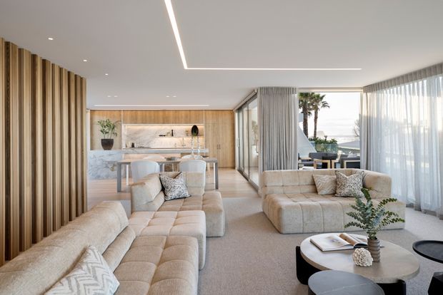 A bright, refined open-plan living space with multiple cream upholstered sofas, a round coffee table, and full-height sheer curtains. A timber-slatted feature wall frames the room, while a marble and timber kitchen with island seating sits behind the living area. Glazing opens toward an outdoor setting with palm trees and water visible beyond.