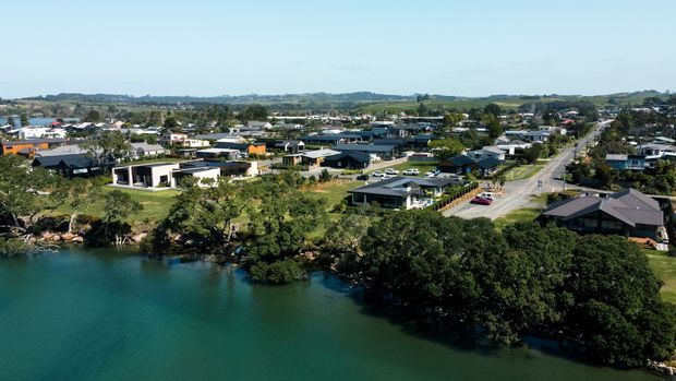 Aerial view of a suburban coastal or waterfront neighbourhood beside calm blue-green water. The area contains detached modern homes, landscaped lots, mature trees, roads, and low rolling countryside in the distance.