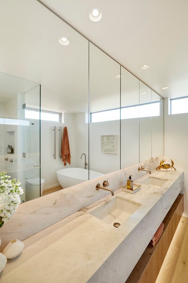 Bright contemporary bathroom featuring a long double vanity with a thick white veined stone benchtop, integrated rectangular basins, timber cabinetry, a freestanding bathtub, glass-enclosed shower, wall-to-wall mirrors, and high horizontal windows. Recessed ceiling lights and warm timber accents create a clean, refined atmosphere.