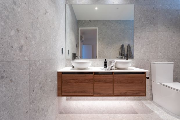 Contemporary bathroom with large-format light grey stone-look wall and floor tiles, a floating dark timber double vanity with white vessel basins, a wide frameless mirror, and a white toilet. Soft under-vanity lighting and recessed ceiling lighting create a clean, refined atmosphere.