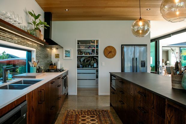 A well-appointed contemporary kitchen viewed from the main work area, with dark timber cabinetry, a large central island, stainless-steel appliances, integrated open shelving, a tiled backsplash, and a warm timber-lined ceiling. Large windows and glazed doors bring in abundant natural light, while globe pendant lights and a patterned runner add character.