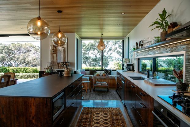 A spacious, open-plan contemporary kitchen with dark timber cabinetry, a large island, pale stone benchtops, and a dark green tiled splashback. The room features a timber-lined ceiling, expansive black-framed windows, layered pendant lighting, open shelving, and views to leafy outdoor surroundings. A dining or living area is visible beyond the kitchen.