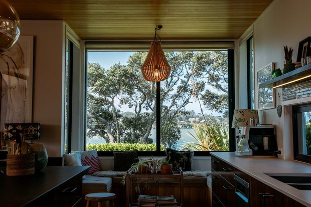 A warm, compact open-plan living and kitchen space with a strong connection to the outdoors. The room features a dark kitchen island in the foreground, dark built-in cabinetry and benchtops along the right wall, a timber-lined ceiling, and a large central window overlooking trees and blue water. A sculptural woven pendant hangs in front of the window, while a low sofa, indoor plants, shelving, and decorative objects create a relaxed, natural interior.