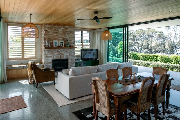 A spacious open-plan living and dining room with a warm timber-lined ceiling, exposed brick fireplace wall, polished concrete or terrazzo-style floor, and expansive black-framed glazing opening to a leafy outdoor setting. A white sofa faces the fireplace and television, while a substantial dark timber dining table is surrounded by woven rattan dining chairs. Ceiling fans, pendant lights, shutters, and layered rugs complete the relaxed coastal-inspired interior.