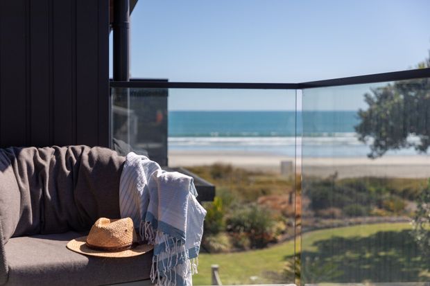 A coastal balcony or terrace overlooking a sandy beach and ocean. The foreground features a dark upholstered outdoor sofa with draped throws and a straw hat, enclosed by frameless glass balustrades. A dark vertical building element and slim roof or balcony structure frame the view.