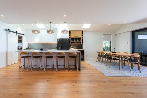 Bright open-plan kitchen and dining area with a large island, five bar stools, white cabinetry, warm timber flooring, pendant lights, and a built-in wine display. A dining table sits beside the kitchen on a textured rug, with sliding doors and windows providing natural light.