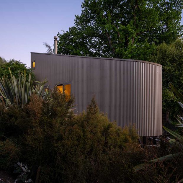 A compact curved-roof or rounded-end building clad in dark vertically ribbed metal, viewed externally at dusk and surrounded by dense native-style planting and mature trees. Small warm-glowing windows and a metal flue provide points of contrast against the muted facade.