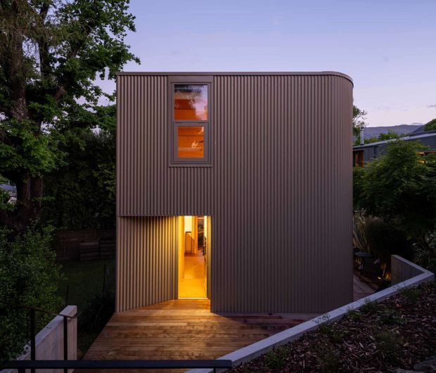 A compact two-storey residential building viewed externally at dusk. The facade is wrapped in vertically ribbed dark metal cladding with a rounded roof-edge corner, narrow upper windows, and a warmly illuminated ground-floor doorway opening onto a timber deck and stair. Dense trees and neighbouring residential buildings surround the structure.
