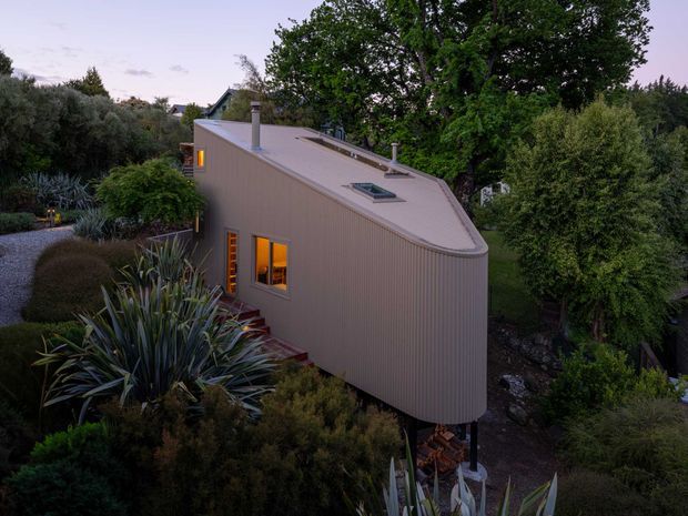 An elevated dusk exterior view of a compact contemporary house nestled within dense landscaped planting. The building has a distinctive rounded-ended, single-storey form with pale vertical cladding, a gently sloping roof, skylights, a chimney, and warm light glowing from narrow windows. Natural stone and timber elements are visible beneath the raised structure, surrounded by mature trees and drought-tolerant garden planting.
