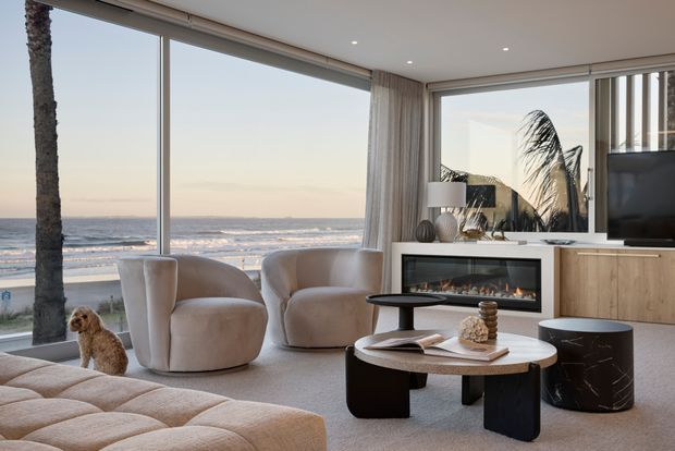 A refined contemporary coastal living room overlooking the ocean, with expansive corner glazing, sheer curtains, curved upholstered lounge chairs, a sculptural coffee table, built-in fireplace, timber cabinetry, and a television. A palm tree and beach are visible outside, while the room is styled in soft neutral tones with rounded forms and a calm, luxurious atmosphere.