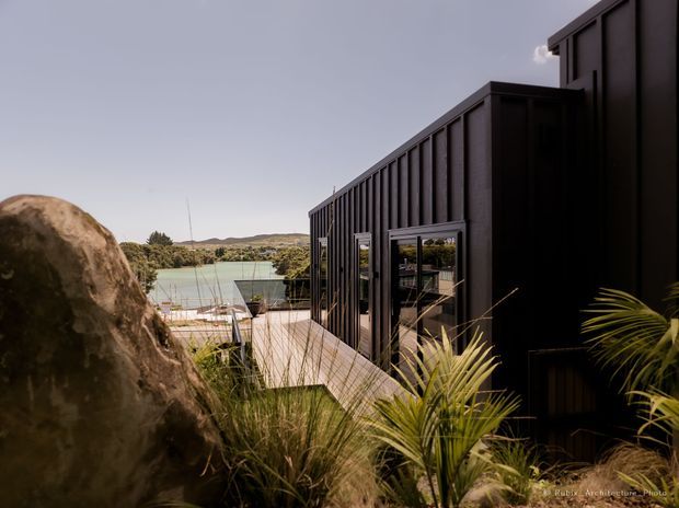 Exterior view of a dark, contemporary single-storey building clad in vertically ribbed black panels, set into a landscaped coastal or rural site. A timber deck and glazed doors lead toward a pale blue-green water body, with native grasses, palms and a large natural boulder in the foreground. The simple rectangular massing and strong vertical facade lines create a minimalist architectural composition.