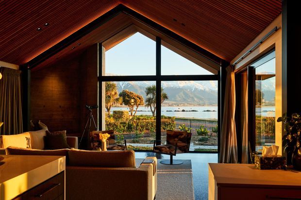 Warm, timber-lined living room with a dramatic pitched ceiling and large gable-end glazing overlooking a lake and mountains. The room contains a sofa seating area, armchair, telescope, curtains, and built-in cabinetry, with strong indoor-outdoor visual connection and sunset-toned lighting.
