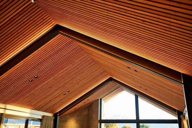 Interior architectural detail showing a dramatic pitched ceiling clad in narrow linear timber battens, with exposed dark structural beams and recessed spotlights. A large triangular clerestory window and floor-to-ceiling glazing bring in daylight, while a concrete-toned wall and curtains are visible below.