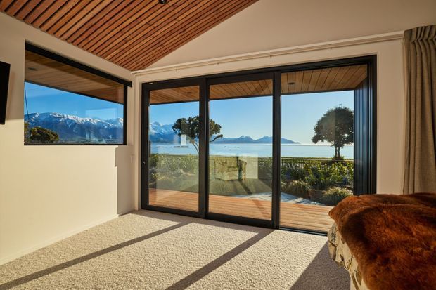 Bright contemporary living space with floor-to-ceiling black-framed sliding glass doors opening to a timber deck and a scenic waterfront view backed by snow-capped mountains. The room has a warm timber slatted ceiling, light neutral walls, soft carpet flooring, full-height curtains, and a plush brown seating piece in the foreground. Strong natural light creates long shadows across the carpet.