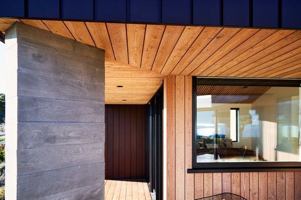A contemporary building exterior with a covered timber-lined entry or veranda. The facade combines warm vertical and horizontal timber boards, a dark horizontal roof or fascia, large black-framed glazing, and a prominent grey concrete wall panel. The timber soffit and decking create a cohesive, warm material palette.