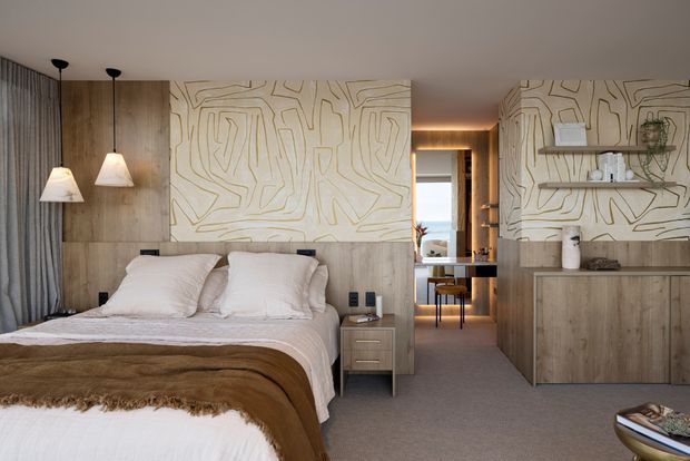 Warm, refined contemporary bedroom with a low upholstered bed, layered neutral bedding, timber wall panelling and built-in cabinetry. A large abstract linear wall treatment spans the headboard wall and continues behind open shelving and storage. Floor-to-ceiling curtains, pendant bedside lights, soft carpet and a view through to a compact desk area create a calm, cohesive interior.