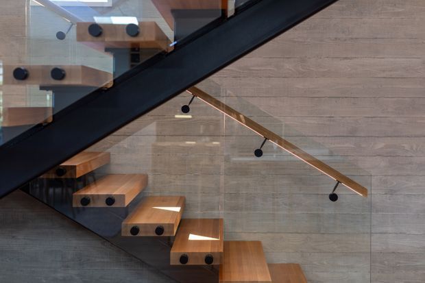 Close-up architectural view of a contemporary stairway with a dark steel or painted metal stringer, timber stair treads, and a glass balustrade. The stair runs diagonally across a textured grey wall, with minimalist black circular light fixtures or fittings suspended along the adjacent timber handrail. Reflections and upper-level steps are visible through the glazing.