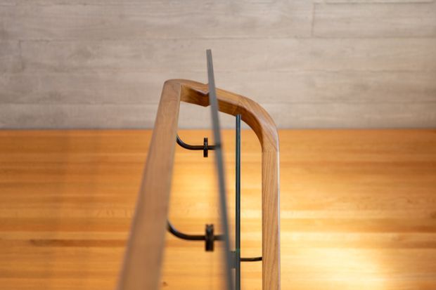 Close-up architectural detail of a timber-framed glass balustrade or stair/void guard, with warm wood flooring and matching pale timber wall panelling. Slim dark metal supports and fittings provide contrast against the natural timber surfaces.