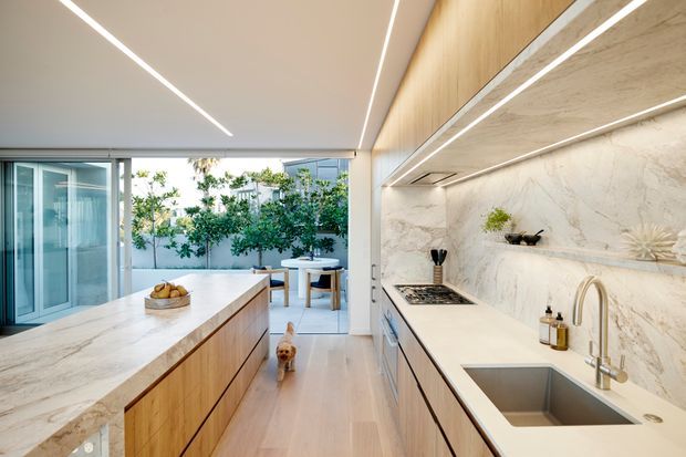 A bright, contemporary galley-style kitchen with a large marble-look island, pale timber cabinetry, integrated linear LED lighting, a stone benchtop and splashback, and a double-bowl undermount sink with brushed metal tapware. Full-height glazing opens toward a small outdoor courtyard or terrace, bringing in abundant natural light.
