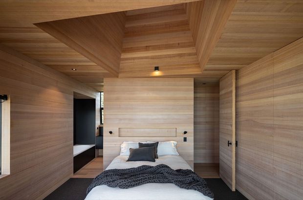 Compact bedroom clad almost entirely in warm natural timber, including the walls, ceiling, built-in headboard, and integrated storage doors. A central bed with white bedding and dark grey cushions and throw sits beneath a recessed timber ceiling detail, with concealed or minimal lighting and dark flooring creating a refined, cosy atmosphere.