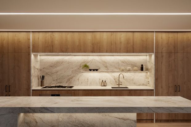 A refined contemporary kitchen featuring full-height warm timber cabinetry, a large pale veined stone island benchtop, and a matching stone splashback with integrated open shelving and concealed lighting. The composition is symmetrical and minimalist, with warm ambient LED lighting and understated fixtures.