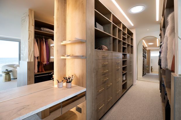 A spacious walk-in wardrobe and dressing area with extensive floor-to-ceiling timber cabinetry, open shelving, drawers, hanging rails, and a central light-wood island with integrated drawers and display shelves. A full-height mirror extends the view down the wardrobe, while a carpeted floor, recessed ceiling lights, and linear LED lighting create a warm, refined dressing space. A bright lounge or bedroom area is visible beyond the opening.
