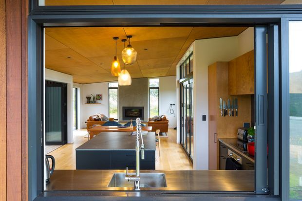 View through a large dark-framed opening into an open-plan kitchen and living area. The kitchen has a broad dark island with an undermount sink and tall brushed-metal tap, timber cabinetry, and warm timber flooring. A timber-lined ceiling, pendant lights, dining table, and lounge seating lead toward a fireplace and full-height glazing. The overall design is warm, refined, and contemporary with strong timber and dark metal elements.