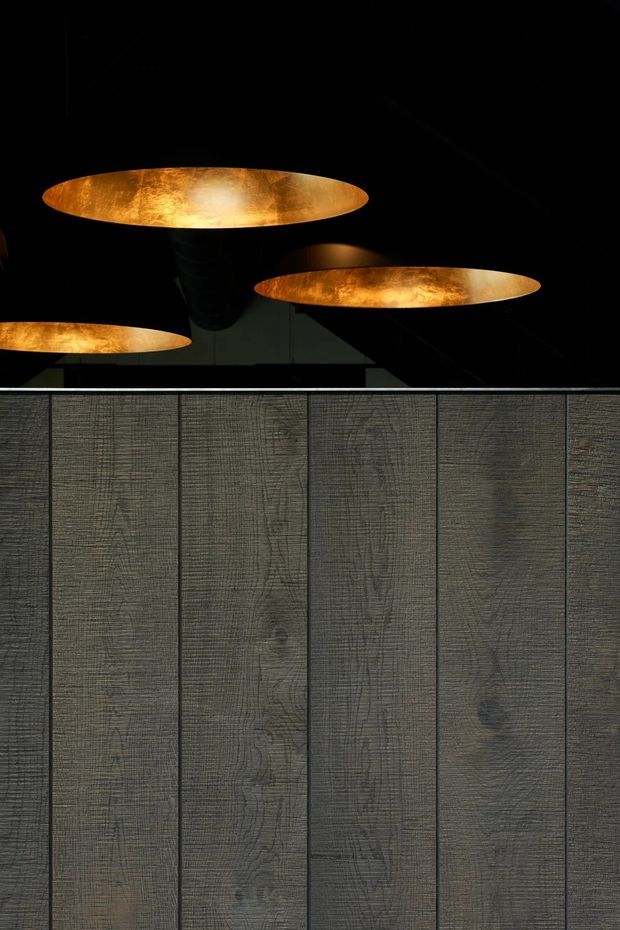 Close-up architectural interior detail showing a dark vertically panelled timber wall or fixed joinery beneath several large, shallow, glowing amber pendant lights. The composition is highly graphic and moody, with warm metallic lighting contrasting against charcoal-black ceilings and grey-brown textured panels.
