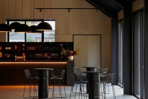 A dark, sophisticated café or hospitality interior with a bar counter and open shelving along the rear wall, tall black-framed windows, high ceilings, and multiple black pendant lights. Small round tables with wire-frame chairs occupy the foreground, while warm under-bar lighting contrasts with timber wall panels and charcoal structural elements.