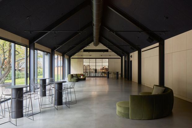 A spacious contemporary commercial interior with an open-plan lounge and hospitality area. The room has a dramatic black pitched ceiling with exposed structural framing and a central metal ventilation duct, full-height glazing along one side, a polished concrete floor, pale wood wall panelling, a distant bar or service counter, and sculptural olive-green lounge seating. High tables and wire-frame stools occupy the glazed edge.