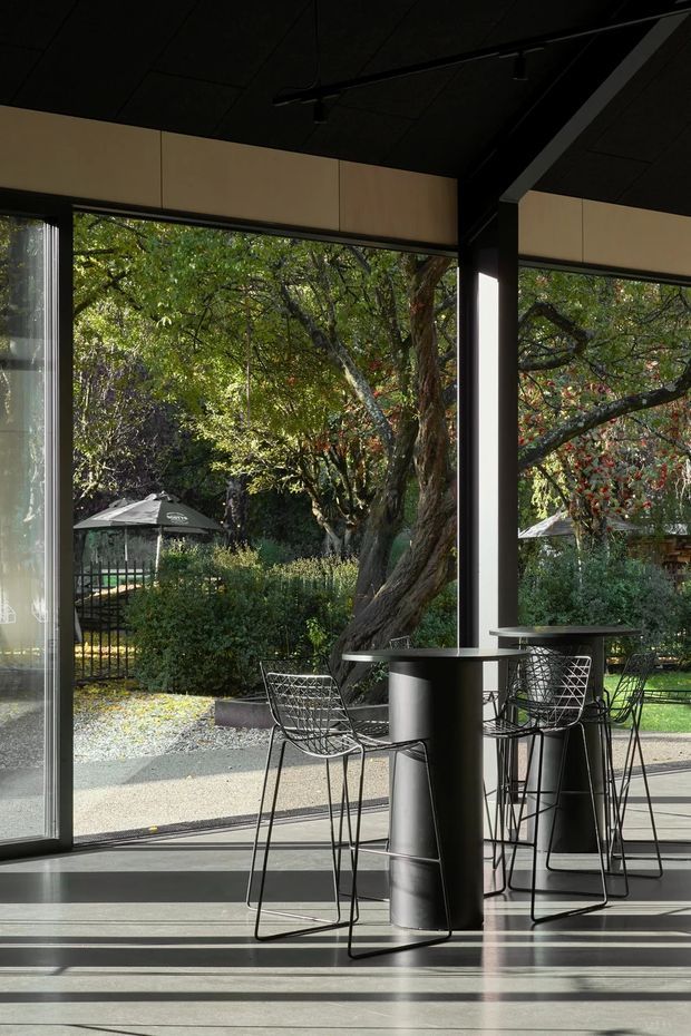 A dark, refined indoor-outdoor entertaining space with full-height glazing opening onto a leafy garden. Black structural columns, exposed dark ceiling panels, slim track lighting, and black wire-frame bar stools surround a tall narrow table. Strong sunlight casts long shadows across the smooth concrete floor, creating a high-contrast, modern pavilion-like atmosphere.