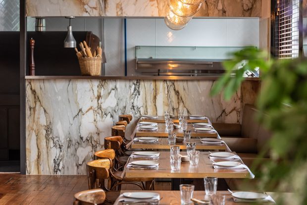 A refined restaurant dining area with a long communal arrangement of timber tables and curved wooden chairs. Dramatic veined marble-clad walls and a matching marble counter frame a built-in service or display area, complemented by dark cabinetry, pendant globe lighting, and warm timber flooring.